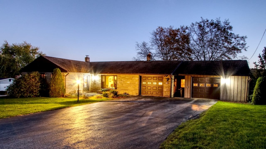 Single-story stone home at dusk with warm exterior lighting, dual garage doors, and lush green lawn.